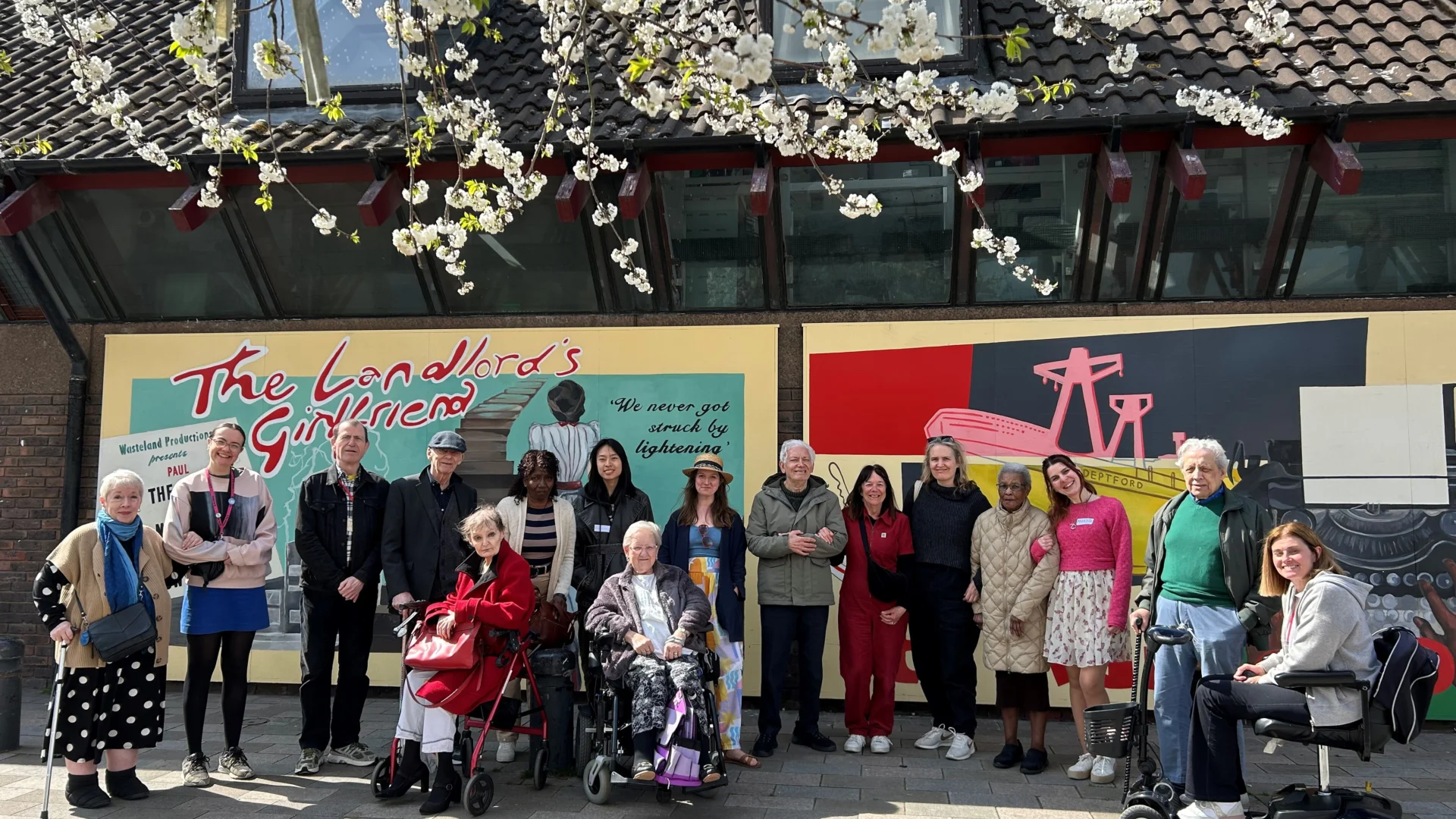 A photo of a group of older people (Meet Me at the Albany members) stood in front of a colourful mural on a sunny day. There are some branches with blossom on them at the top of the frame.