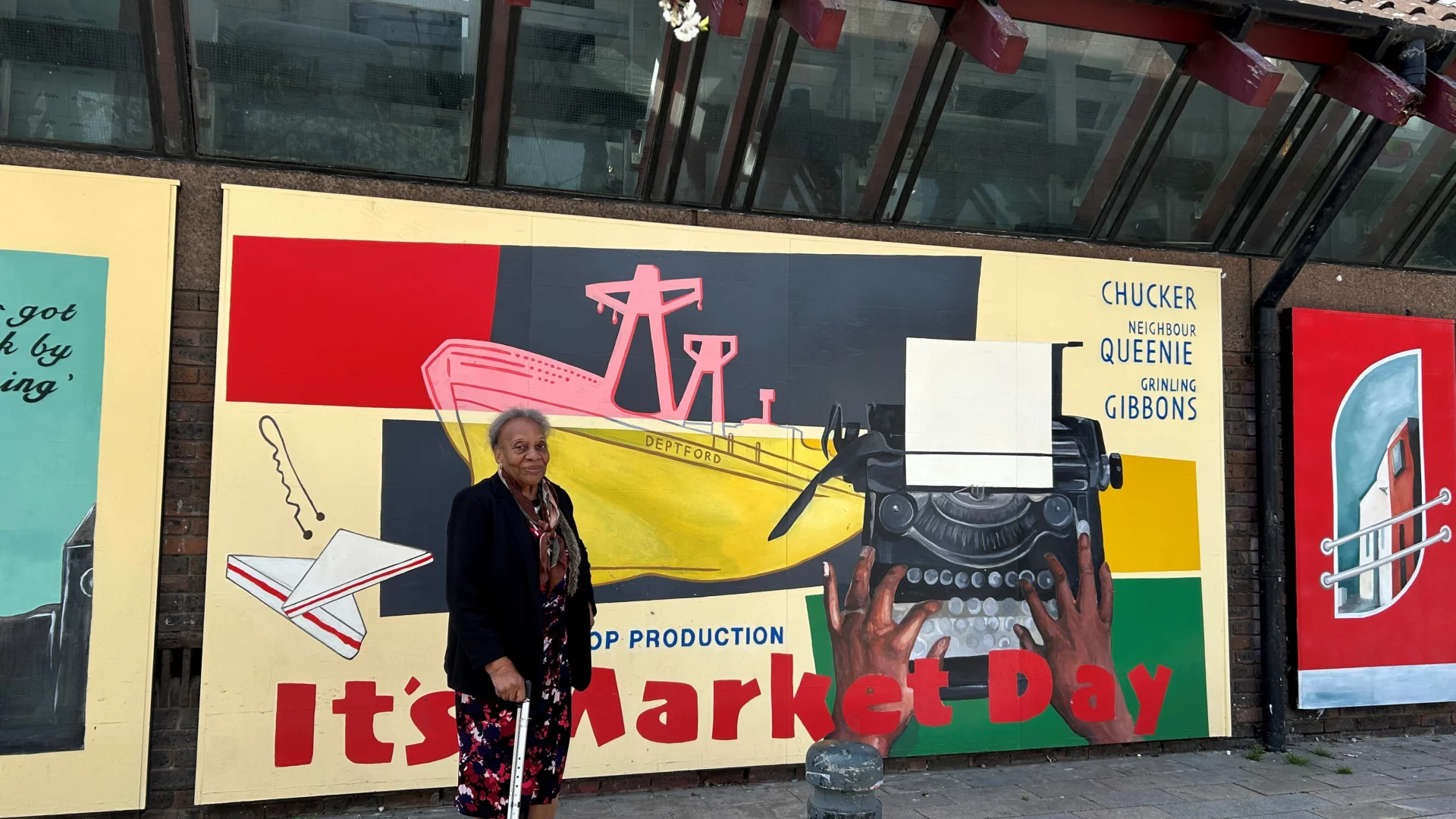 A black woman holding a walking stick stands in front of a brightly coloured mural. The mural has red text saying 'It's market Day' and there are illustrations of a ship, a sandwich and a typewriter.