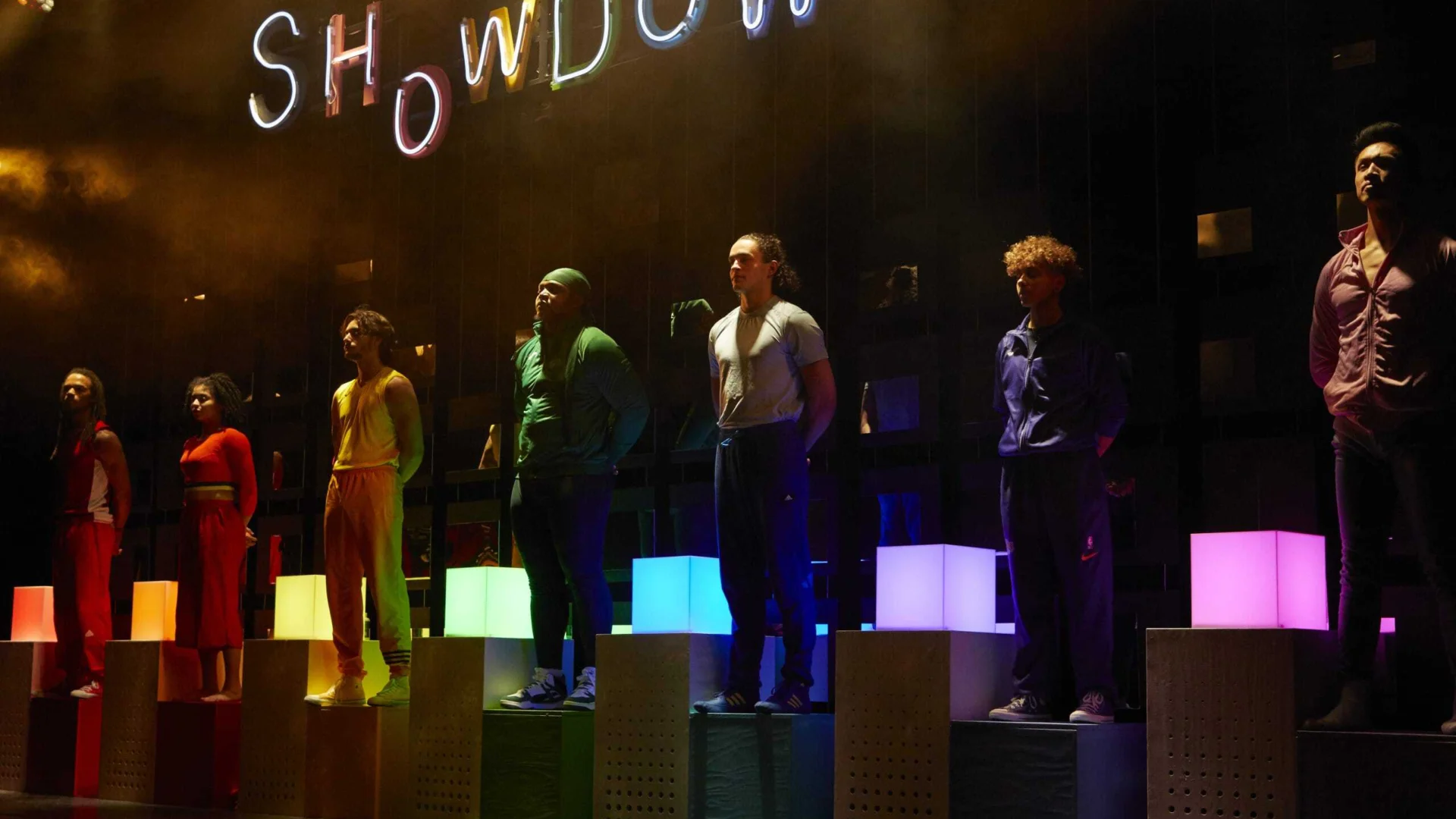 A photo of seven performers, each wearing a different bright colour and stood on a light up plinth in the same colour that they are wearing. Behind them the word 'Showdown' is lit in rainbow colours.