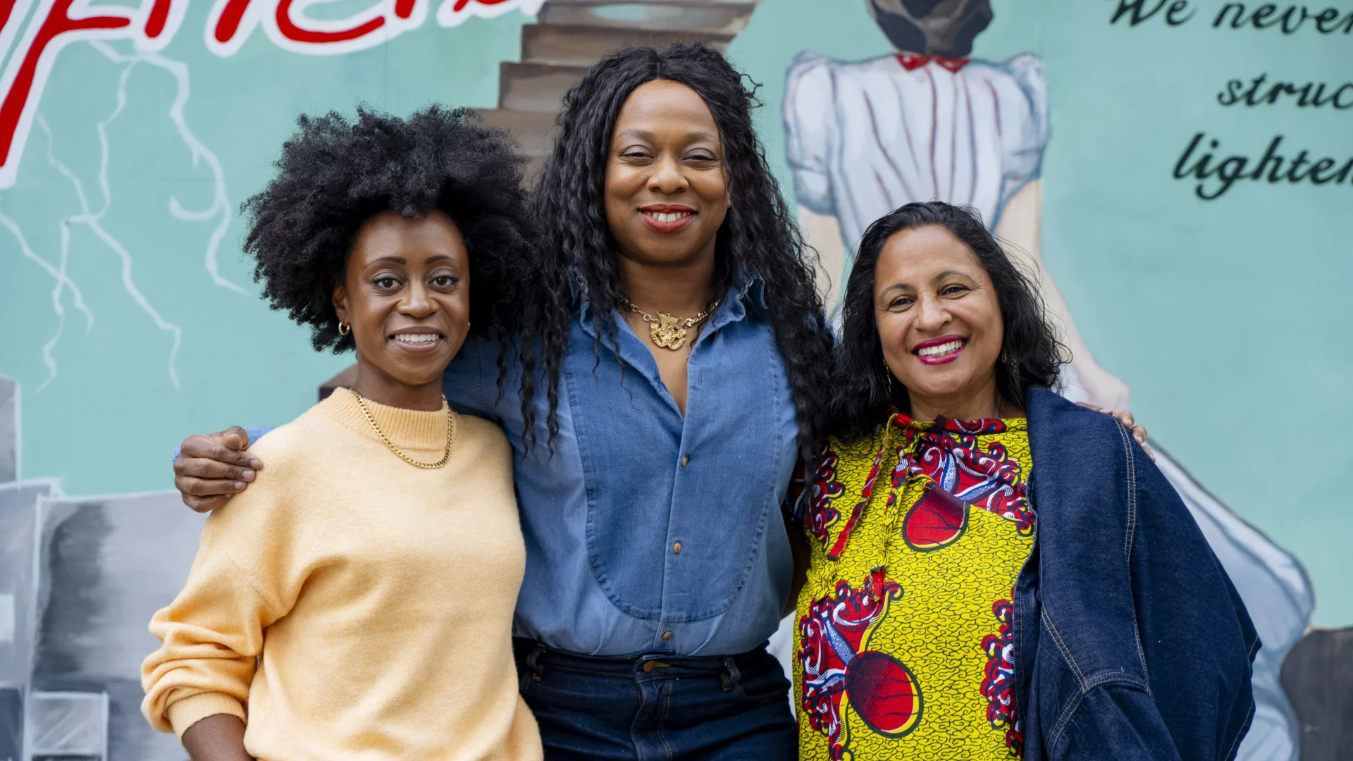 Albany Exec Director Mimi Findlay, Creative Director Vicki Amedume, and Chair of the Board Michelle Matherson, standing together in front of a colourful blue mural.