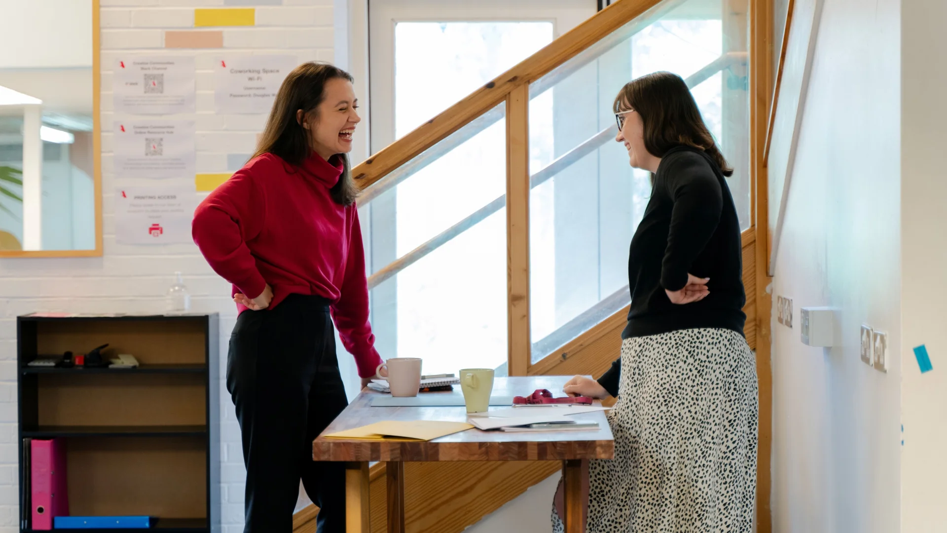 Two women with straight brown hair stand on either side of a low table, chatting and laughing.