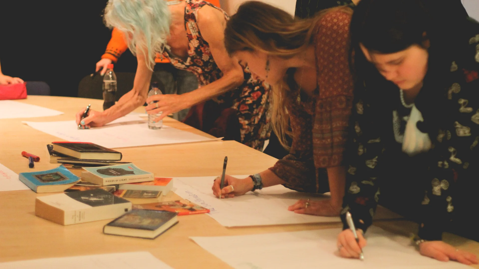A photo of three women stood over a table, writing on large sheets of white paper. In the middle of the table are pens and a pile of books.