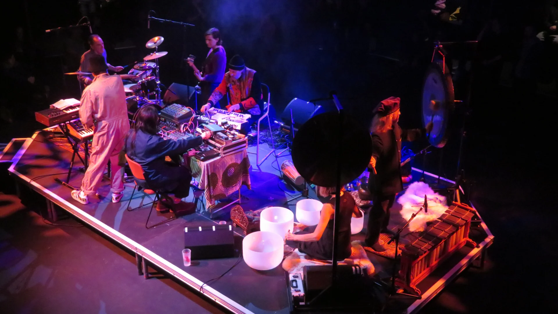 A photo of a stage from above. On the stage are musicians playing the drums, keyboards and guitar. They are lit in pink and purple lighting.
