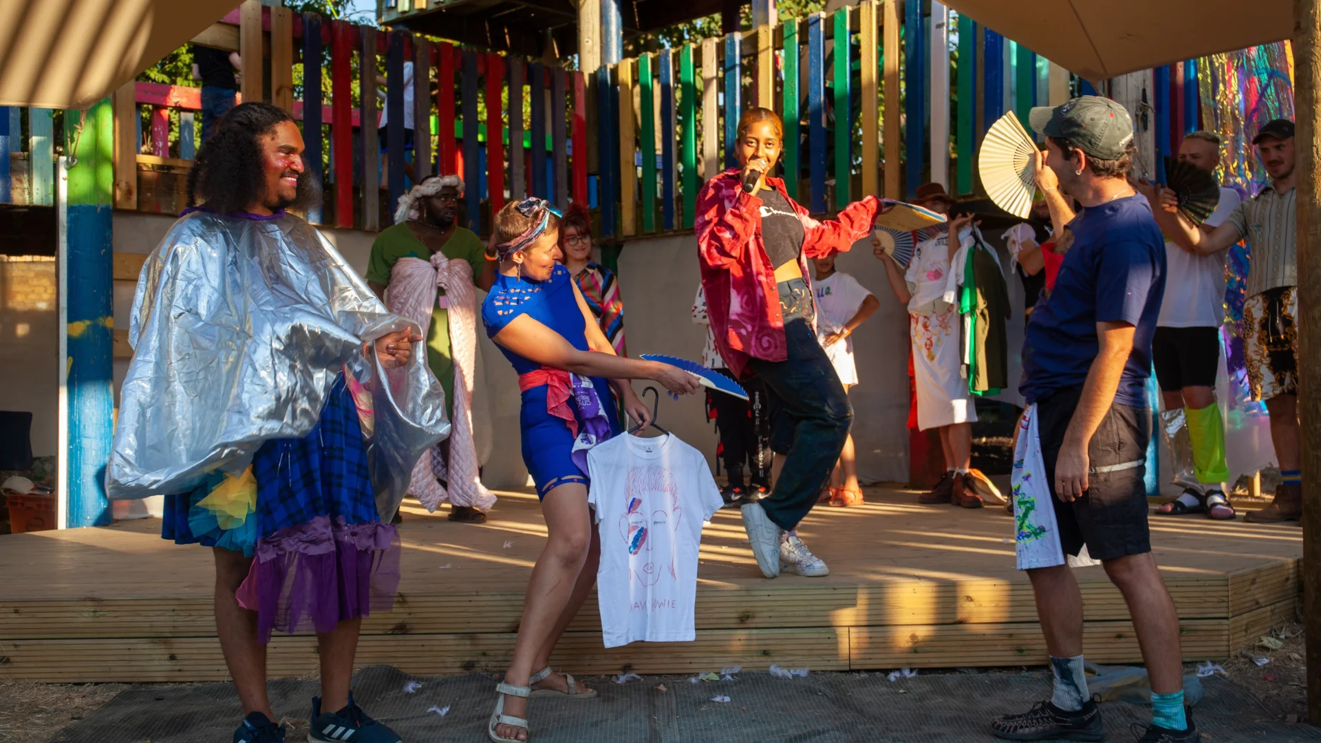 A photo of a group of people wearing bright colours on a stage on a hot, sunny day. A young black woman wearing a red bomber jacket is speaking into a microphone. Surrounding her are people wearing bright outfits and waving paper fans.