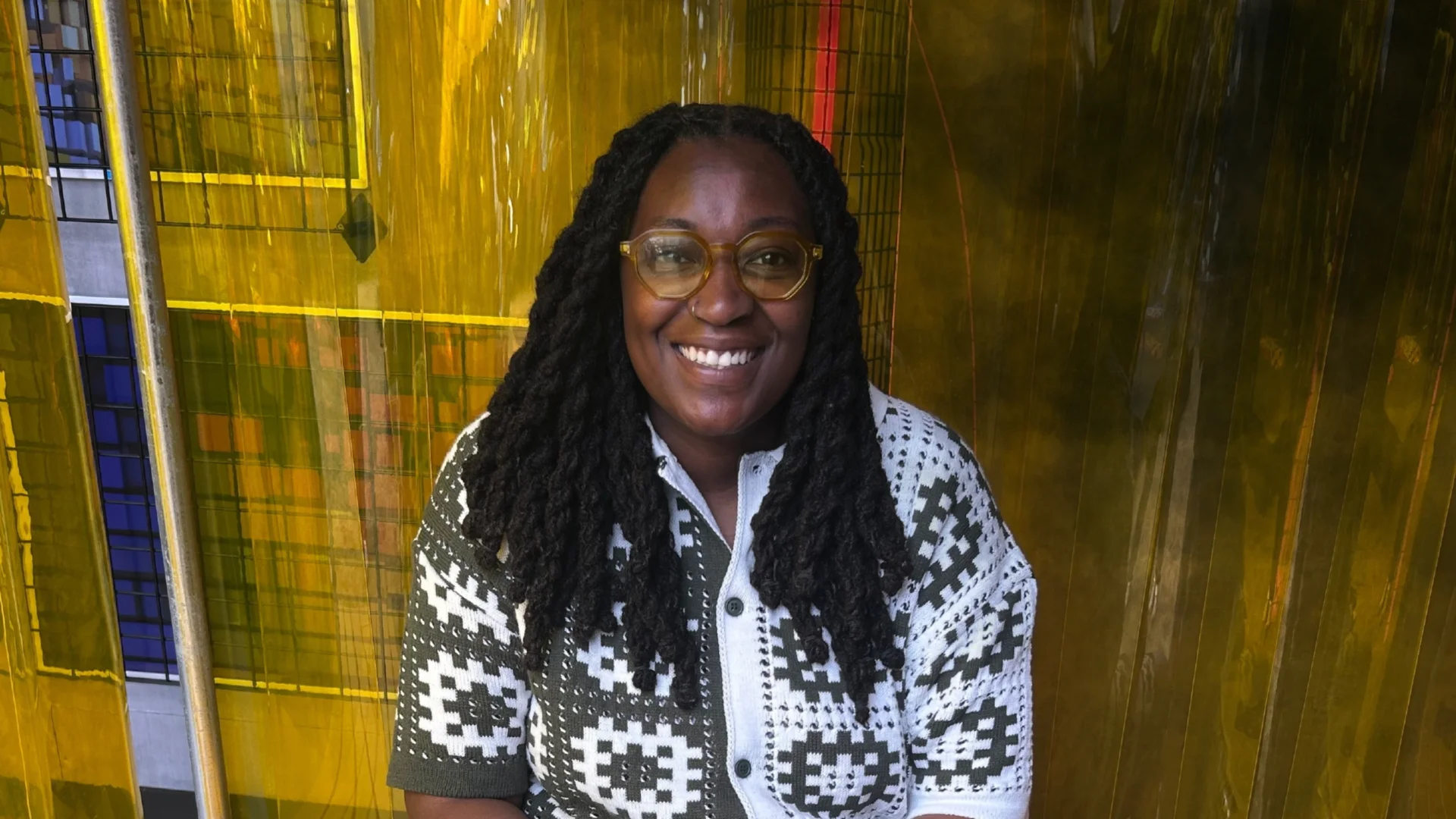 A photo of a black woman with long braids, wearing a white and grey crochet top, sat in front of a yellow wall and smiling at the camera.