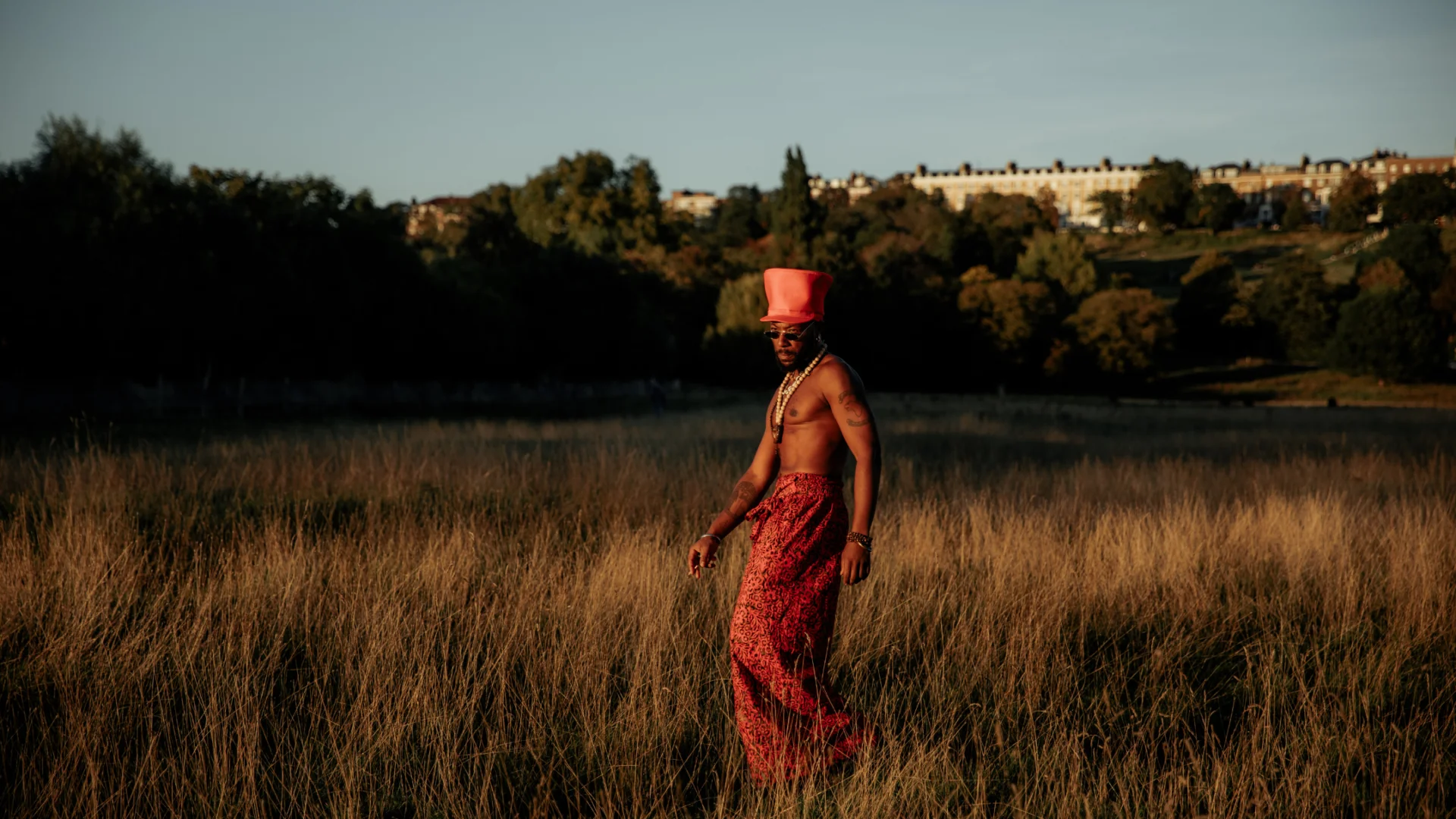 A photo of a black man wearing red trousers and a red top hat, walking through a field of long grass on a sunny day.