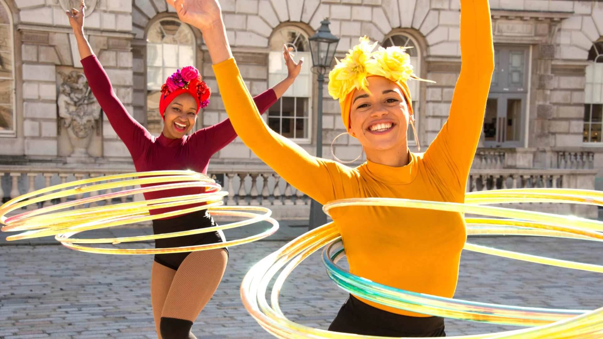 A photo of two women hula hooping in the square of Somerset House. They are wearing bright colours and headscarves and are smiling.