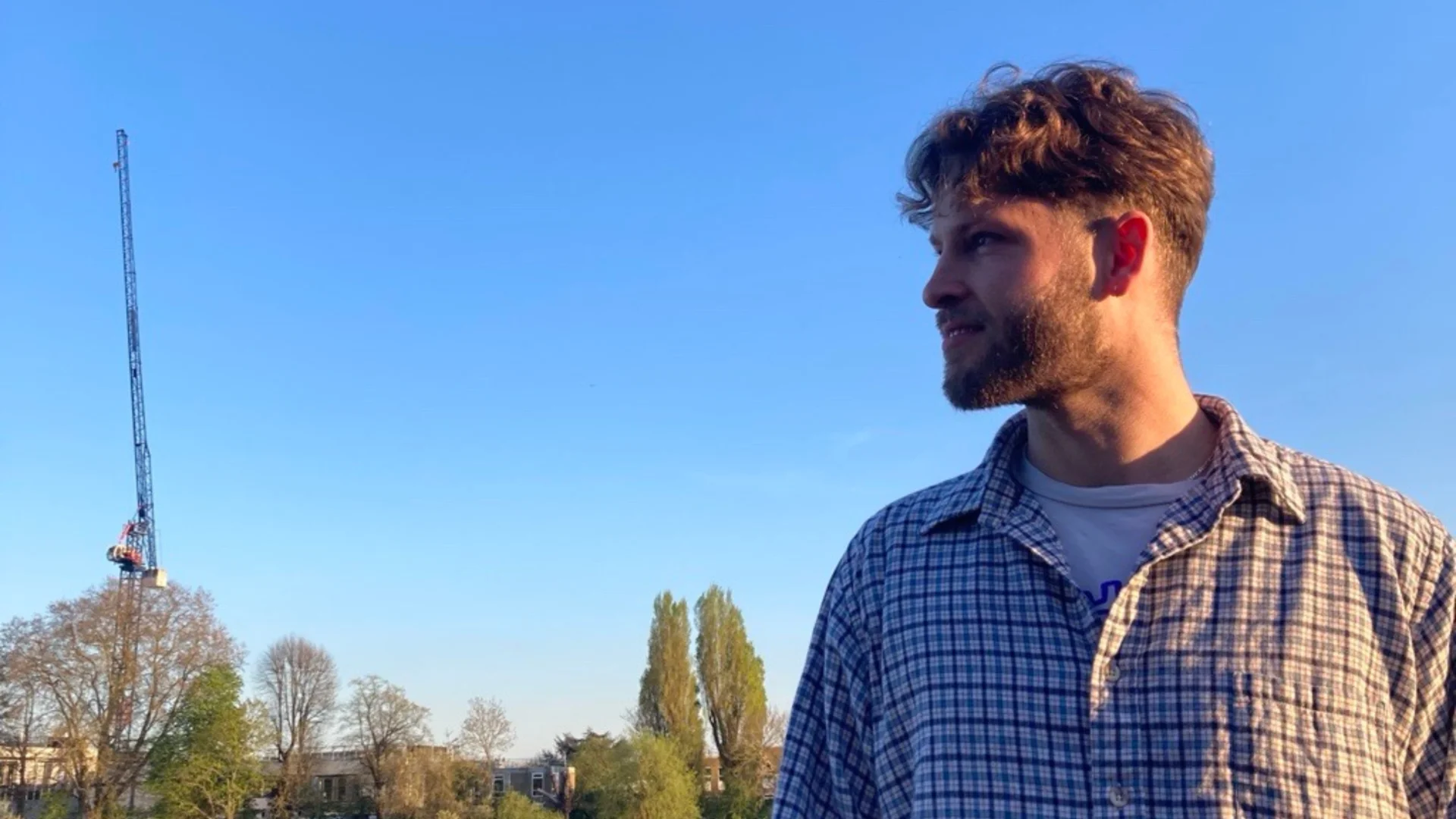 A photo of a white man wearing a checked shirt, gazing to the left. He is stood outside with trees and a crane in the background.