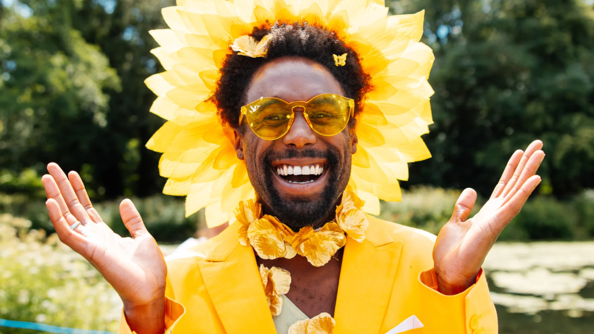 A photo of a black man dressed all in yellow, with yellow sunglasses and a yellow headdress which looks like a sun. He is smiling broadly with his arms open wide.