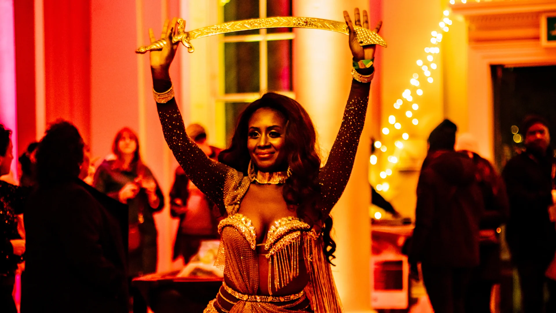 A photo of a black woman wearing a gold glittery dress and holding a gold sword raised above her head. She appears to be at a party, the room is lit with pink and yellow light and there are people mingling behind her.