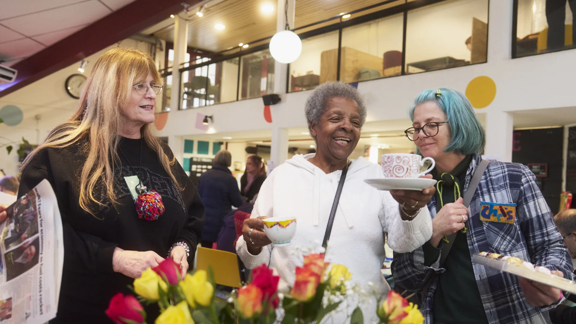 A photograph of three women chatting and holding cups of tea in the Albany cafe. In front of them is a large bunch of yellow and pink flowers.