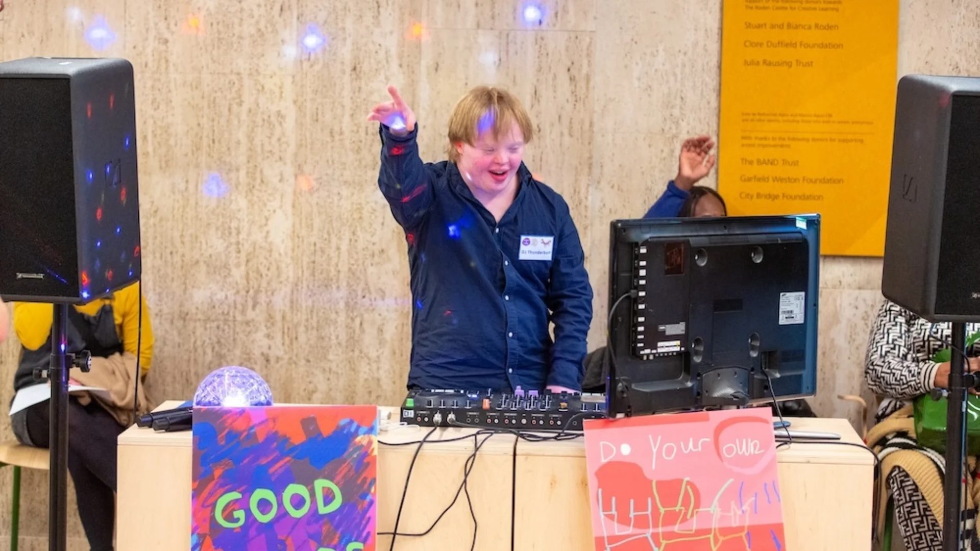 A photo of what a white man wearing overalls, smiling with his arm raised in the air, stood behind a DJ desk. Two colourful signs are in front of the table which read 'Good chaos' and 'Do Your Own Thing'.