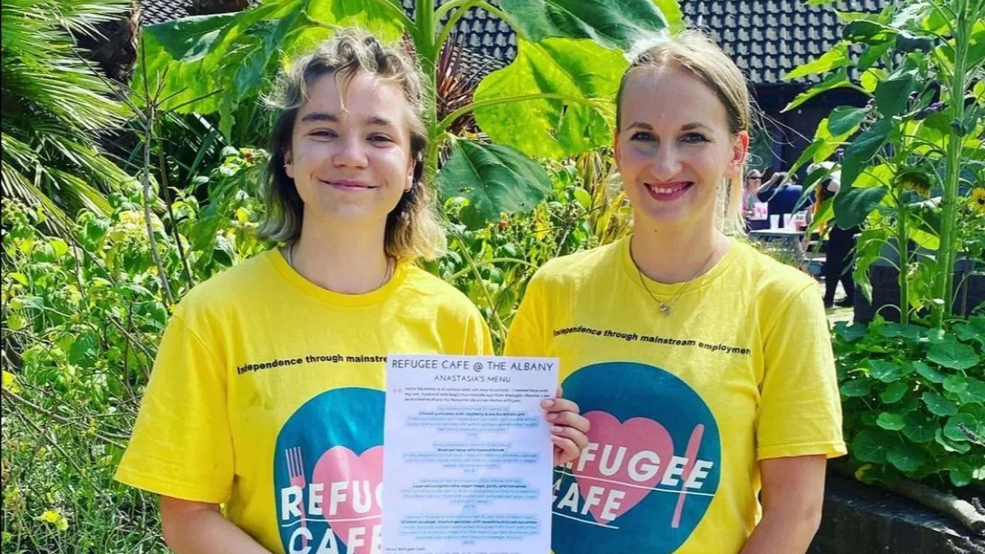 A photo of two white women wearing yellow t-shirts with blue and pink 'Refugee Cafe' logos on them. They are stood smiling in the Albany garden and are holding a cafe menu.