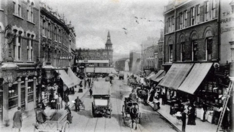 A black and white photograph of a street in Deptford in the 1800s, with shops on either side and horse and carriages driving down the road.