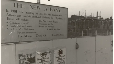 A black and white photo of hoardings surrounding a building site. Text on the hoarding reads: The New Albany: In 1981 the building on this site will rehouse the new Albany and provide additional facilities for Deptford. These will include: A children's centre, A family advice centre, Arts and crafts workshops, Restaurant and bar, rooms for meetings and social activities.'