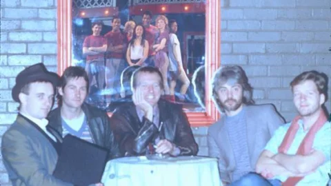 A faded photograph of five men (the band Squeeze) sat around a fold out table and chairs in the foyer of the Albany.