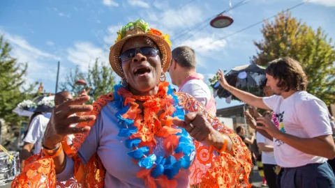 A photograph of a black woman wearing bright orange and blue feather boas. She is outside in the middle of a carnival or similar event and is looking at the camera and singing.