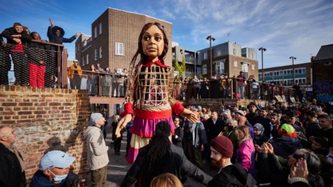 A giant puppet of a young girl, with long brown hair and wearing a red, gold and pink dress, stands amongst a crowd on a sunny day in Deptford Market Yard.