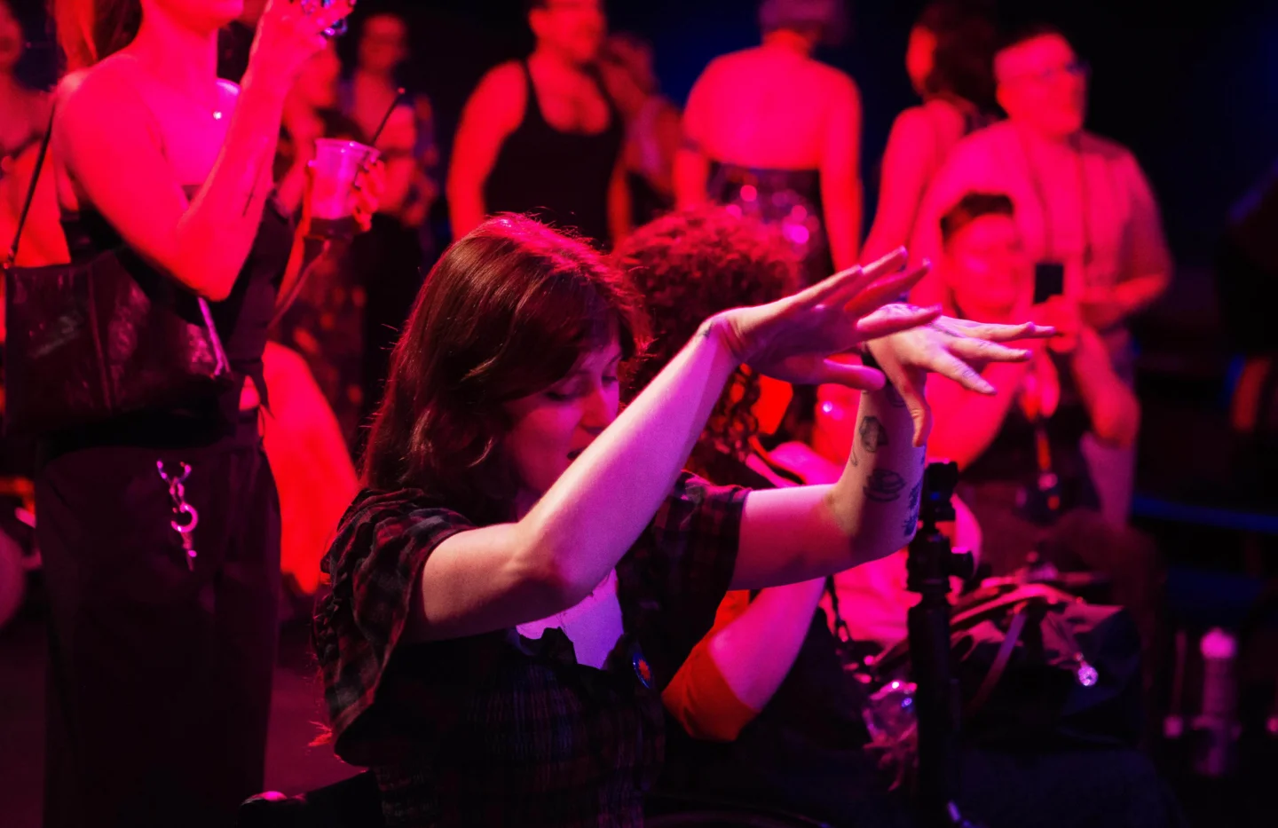 A photo of two people in wheelchairs dancing with their arms in the air. They are bathed in pink light.