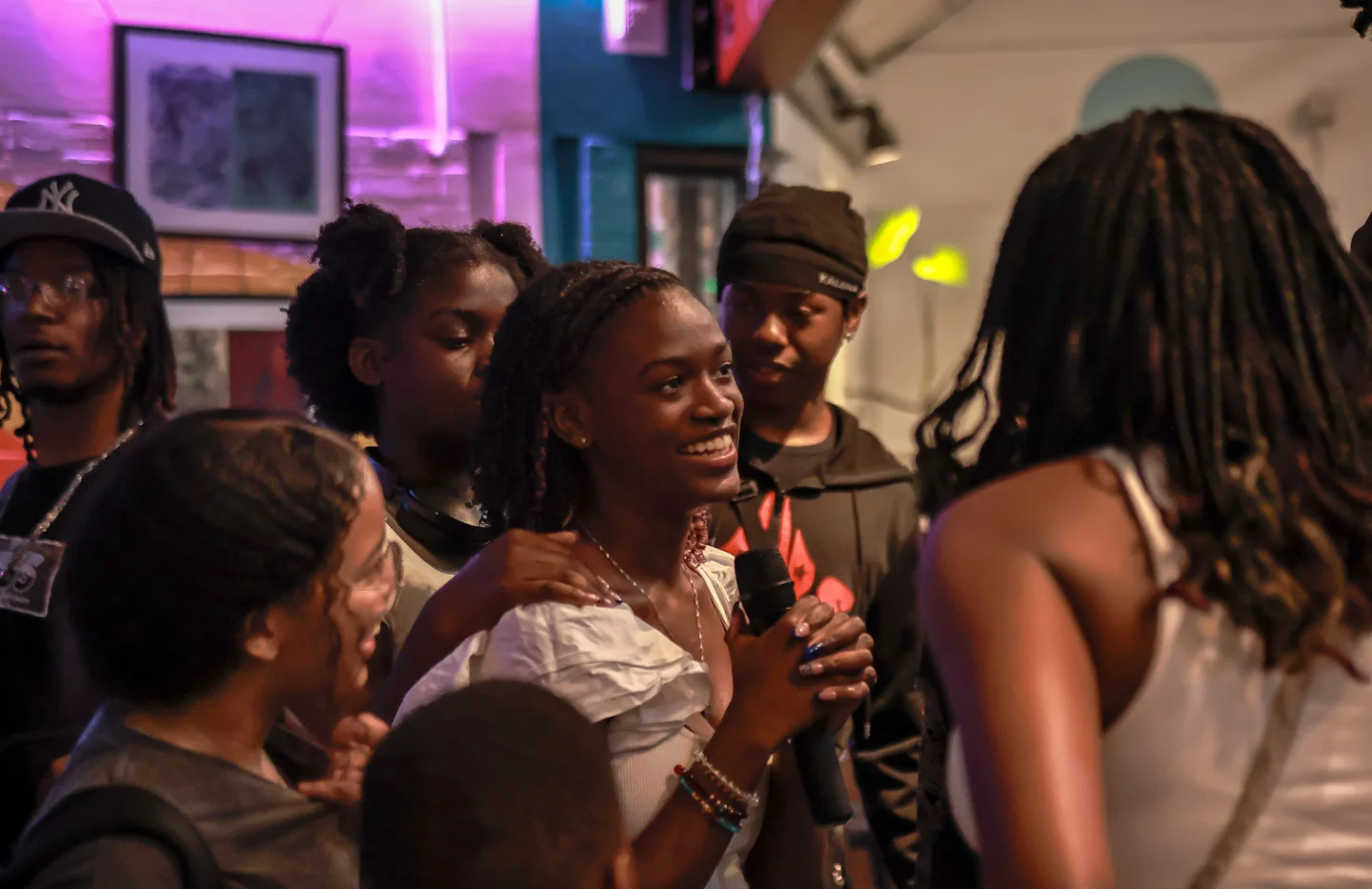 A group of young people gathered in a lively indoor space, with one girl in the centre smiling and holding a microphone.