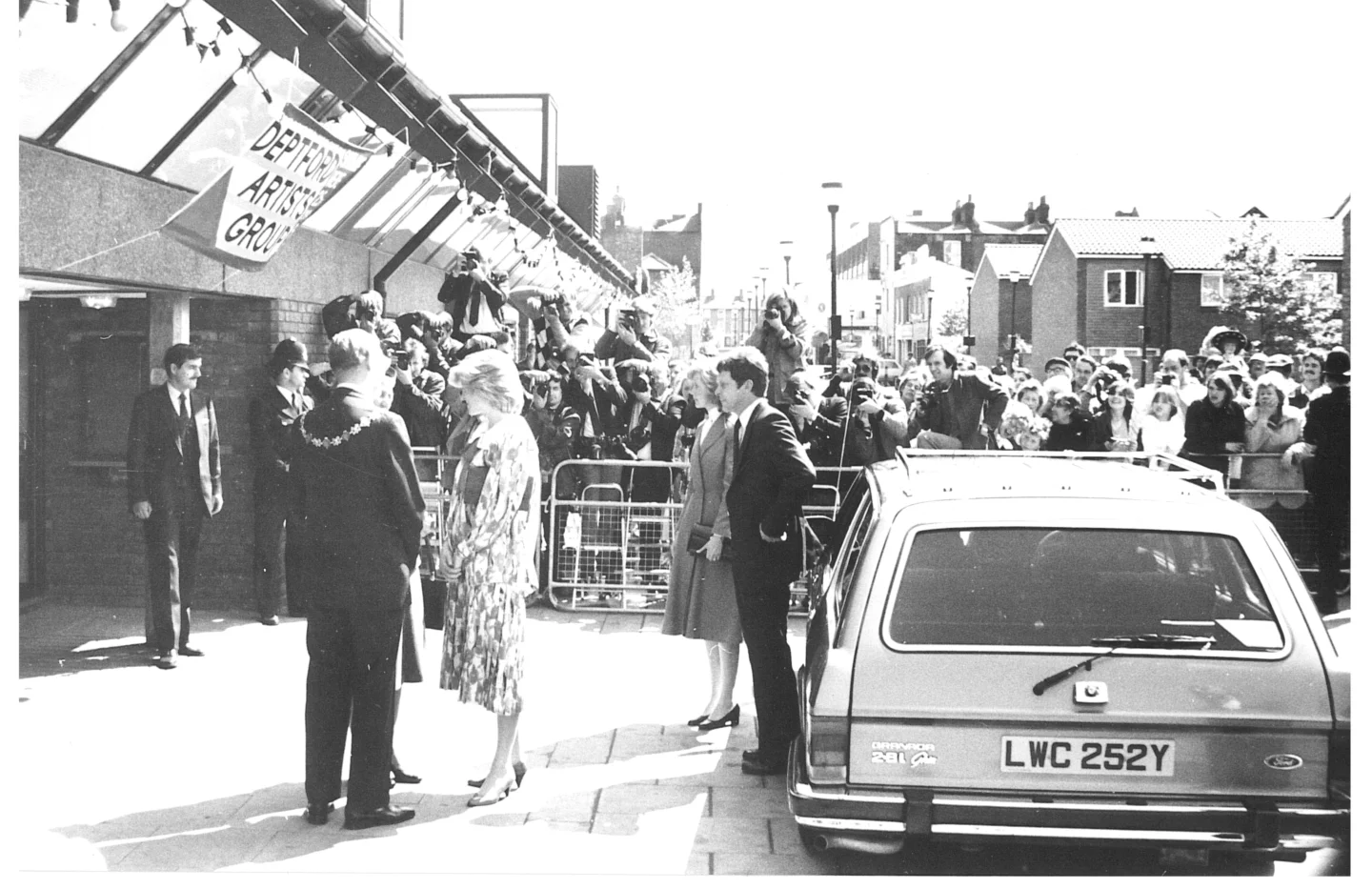 A black and white photograph of Princess Diana having just stepped out of a car at the entrance to the Albany. Behind her there is a crowd watching on.