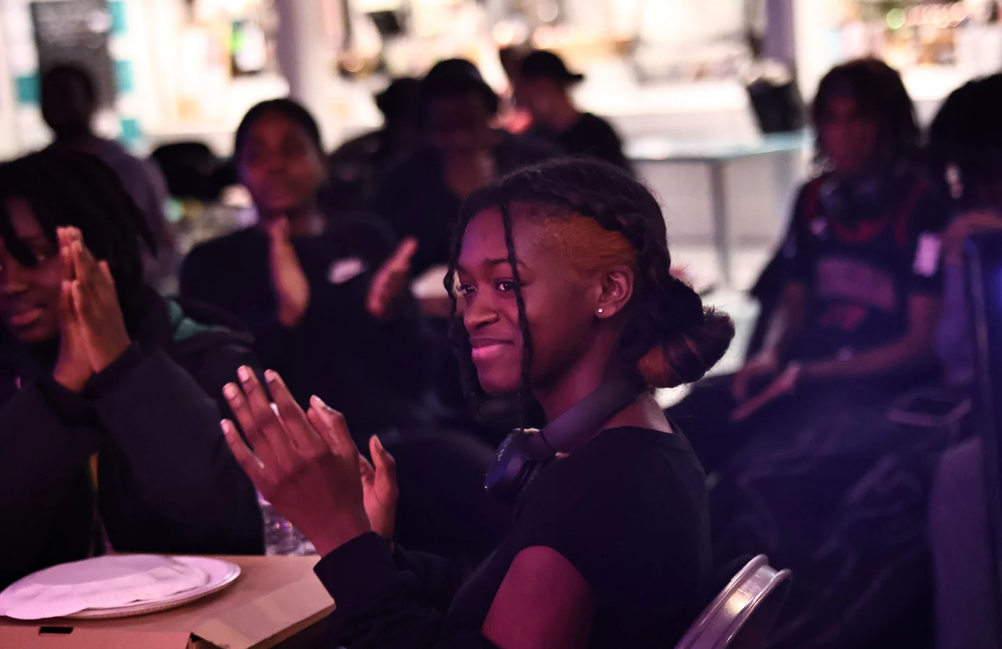 A photo of a young black girls sat in a crowd. She is wearing a black hoodie, looking off to one side and smiling and clapping.