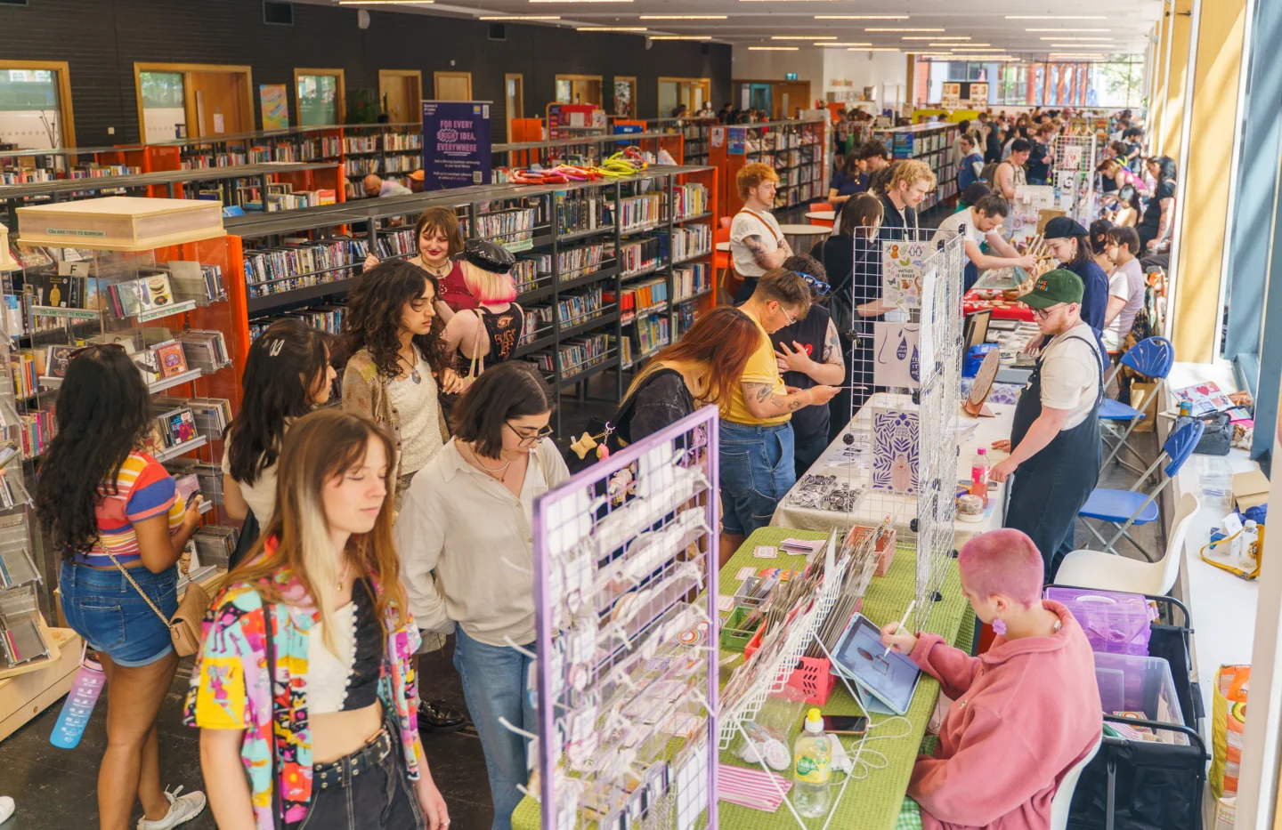 A row of craft stalls in the library at Deptford Lounge. The stalls are parallel to the bookshelves in the library and people are milling around, chatting to the stallholders.