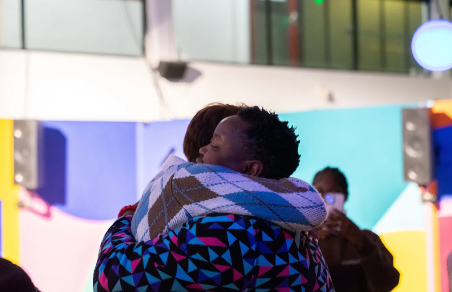 Two people embracing in a warm hug. They are in a colourful, vibrant indoor space with soft lighting.