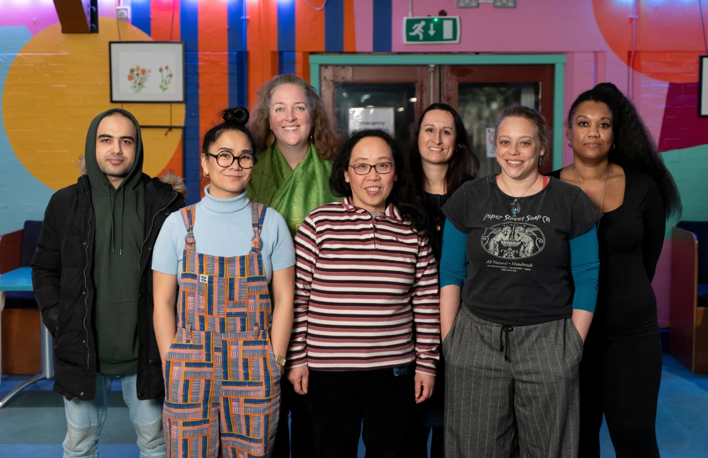 Seven people stand indoors in a row, posing for a group photo against a colorful mural background.