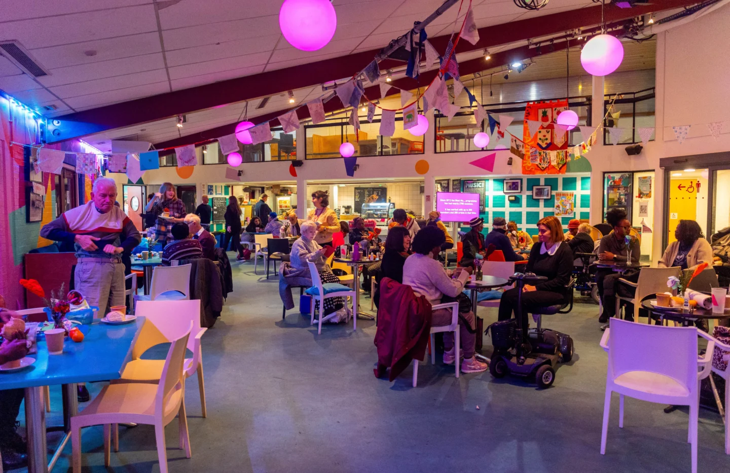 A photo of the Albany cafe during a Meet Me party. The tables are full of people chatting, there is bunting hanging from the ceiling and pink lighting.