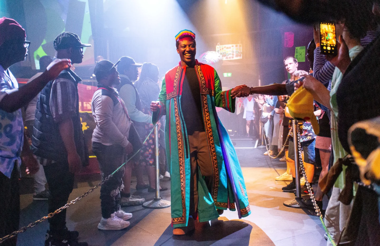 A photo of a young black man wearing bright colours on a catwalk. There is brightly coloured lighting and people on either side of the catwalk are cheering him on.