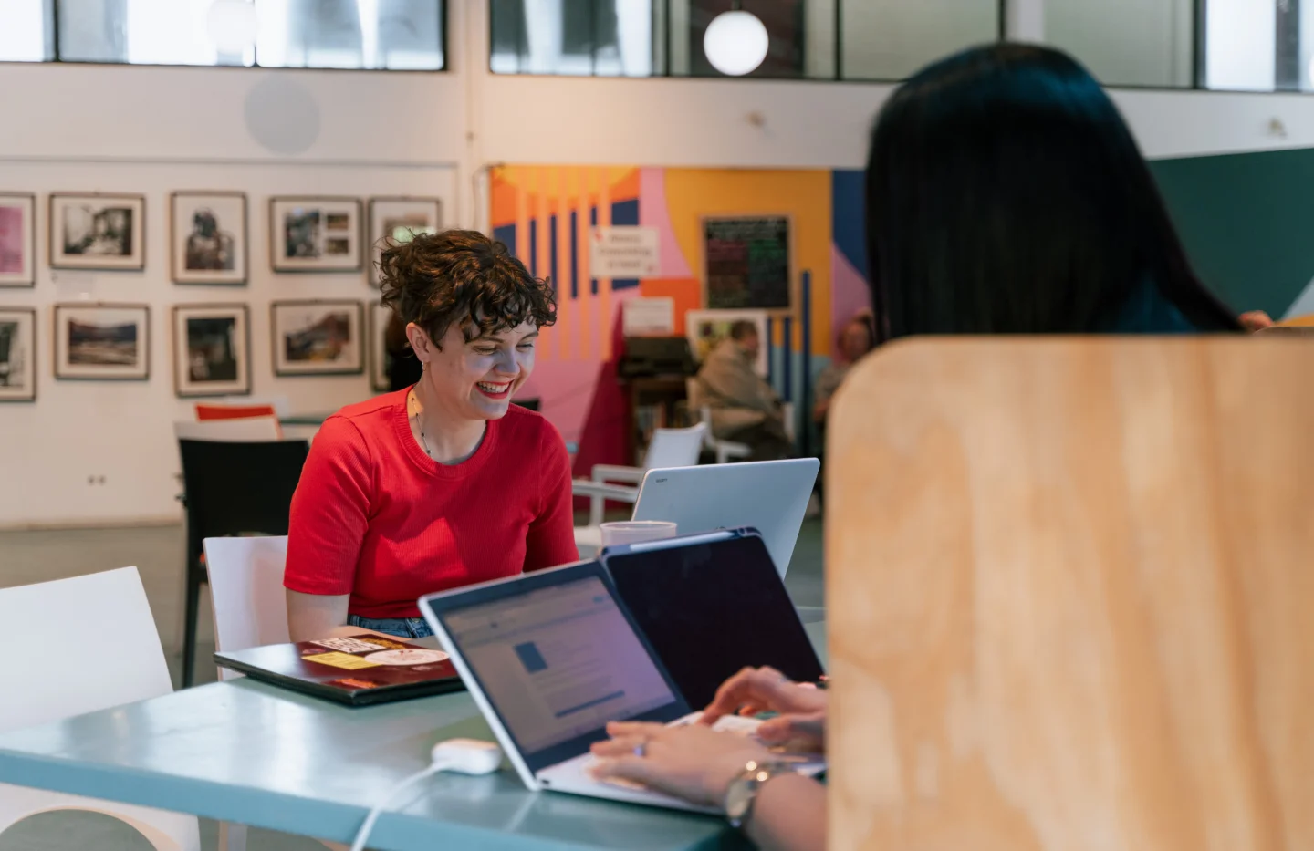 A photo of two people sat at a table on laptops. One has their back to the camera. The other is a white woman with a red top and curly hair, she is smiling.