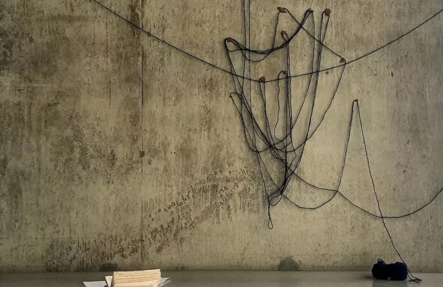 A photo of an artwork. Some black string is hung up on a grey concrete wall and there are papers spread out on the floor.