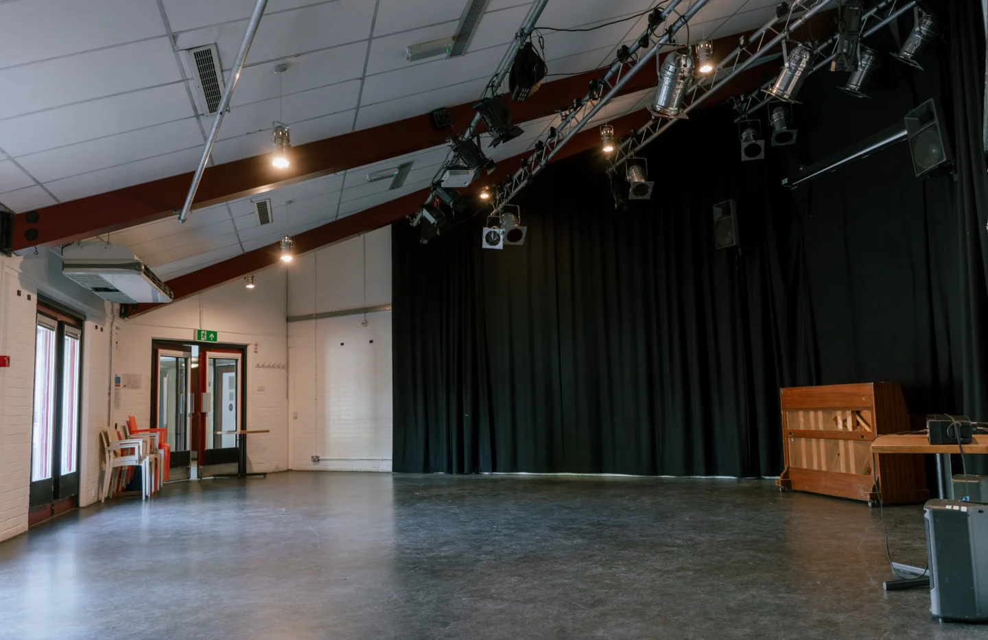 A photo of the Red Room at the Albany. It's an empty space with a lighting rig on the ceiling, and black curtains surrounding the walls. There is a piano in the corner.