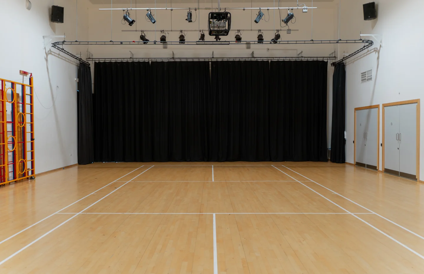 A photo of the Main Hall at Deptford Lounge. It looks like a school sports hall, with court lines on the floor, and gymnastics equipment along one wall. There is a lighting rig on the ceiling and a black curtain covering the back wall.