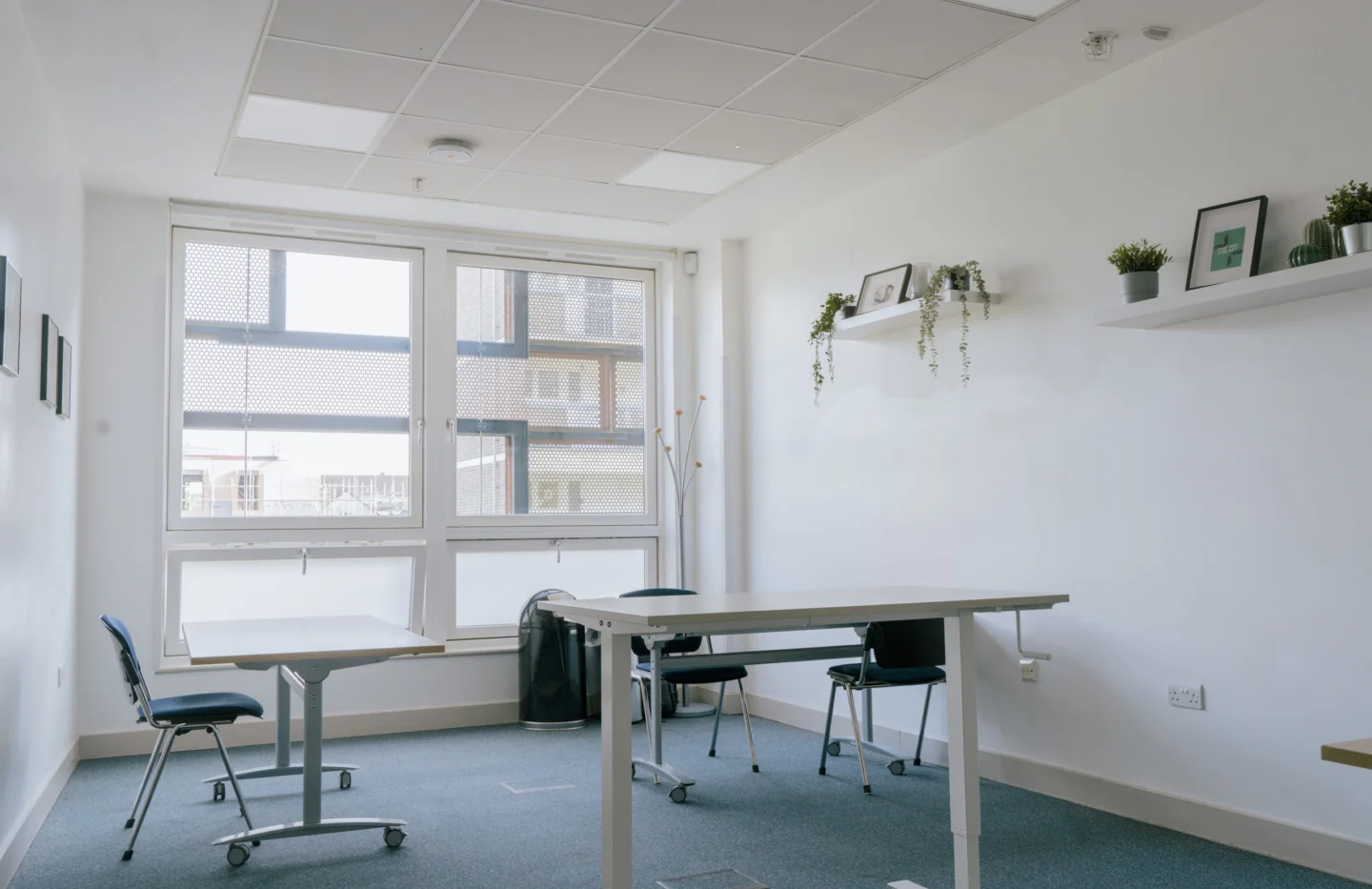 A photo of a meeting room with white walls and one wall is a floor to ceiling window. Inside there are two desks, chairs and there are plants and photo frames on the shelves on the walls.