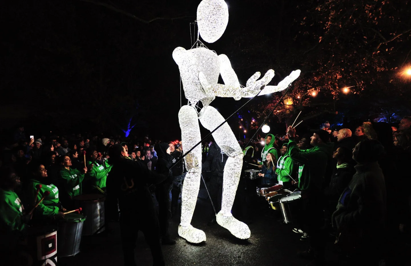 A photo of a giant human puppet lit up in bright white light. It is nighttime and through the dark you can just glimpse people in green t-shirts surrounding the puppet and drumming, and crowds of onlookers behind them.