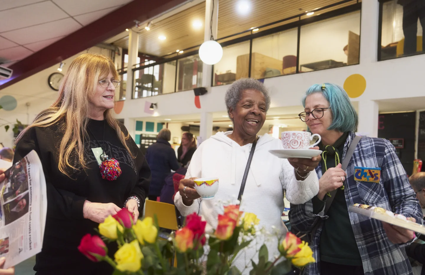 A photograph of three women chatting and holding cups of tea in the Albany cafe. In front of them is a large bunch of yellow and pink flowers.