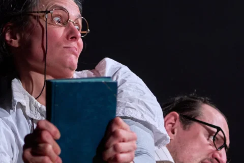 A photo of a woman wearing round glasses and a white shirt clutching a book. Behind her a man wearing glasses leans down - you can just see his head at the bottom of the photo.