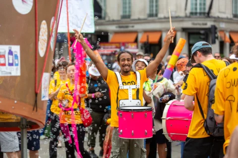A photos of a band wearing yellow t-shirts on the street. A man in the centre of the tshirt is playing a pink drum and has his hands in the air