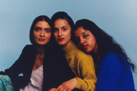 A posed photograph of three Asian women sat closely together and gazing into the theatre. They all have long dark hair and the backdrop behind them is pale blue.