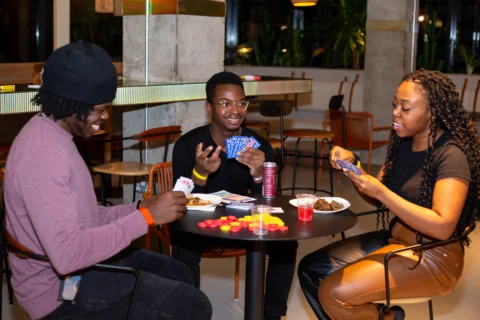 A photo of three people, two black men and one black woman, sat around a table playing cards and eating snacks.