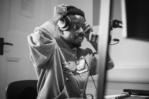 A black and white photo of a young black man wearing a hoodie, holding headphones over his ears.