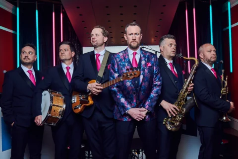 A group of musicians dressed in suits with bright pink ties stand in a line holding their instruments.