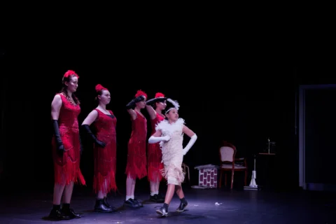 A photo of a group of young performers on stage. They are dressed in 1920s dresses and feather hats. There is a line of performers in red dresses and one in white struts in front of them.