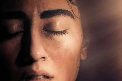 A closeup photo of a presumed woman's face with eyes closed, her left side is in darkness, but light pours in from the top right corner, juxtaposing light against the shadows of what lies in the darkness.
