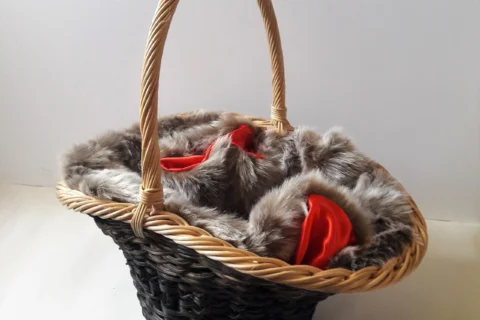 A woven with a darker material body and a light wicker handle is filled with grey fur lined with a red fabric. The fur and fabric mix is mixed in the basket so it all seems to be a part of the same item. The basket is set on a white background.