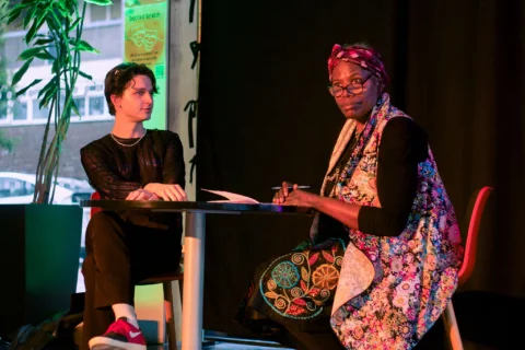 A photo of a young white man and a black woman wearing a headscarf and colourful cardigan sat at a table. Behind them is a black curtain and a pot plant.