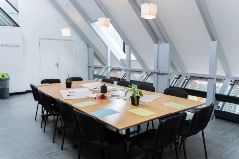 A photo of a meeting room with an empty table and chairs in the middle, set for a meeting with papers, pens and pot plants.