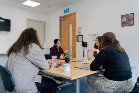 A group of women sit around a square table having a conversation. On the table there are mugs, workbooks and a pot plant.